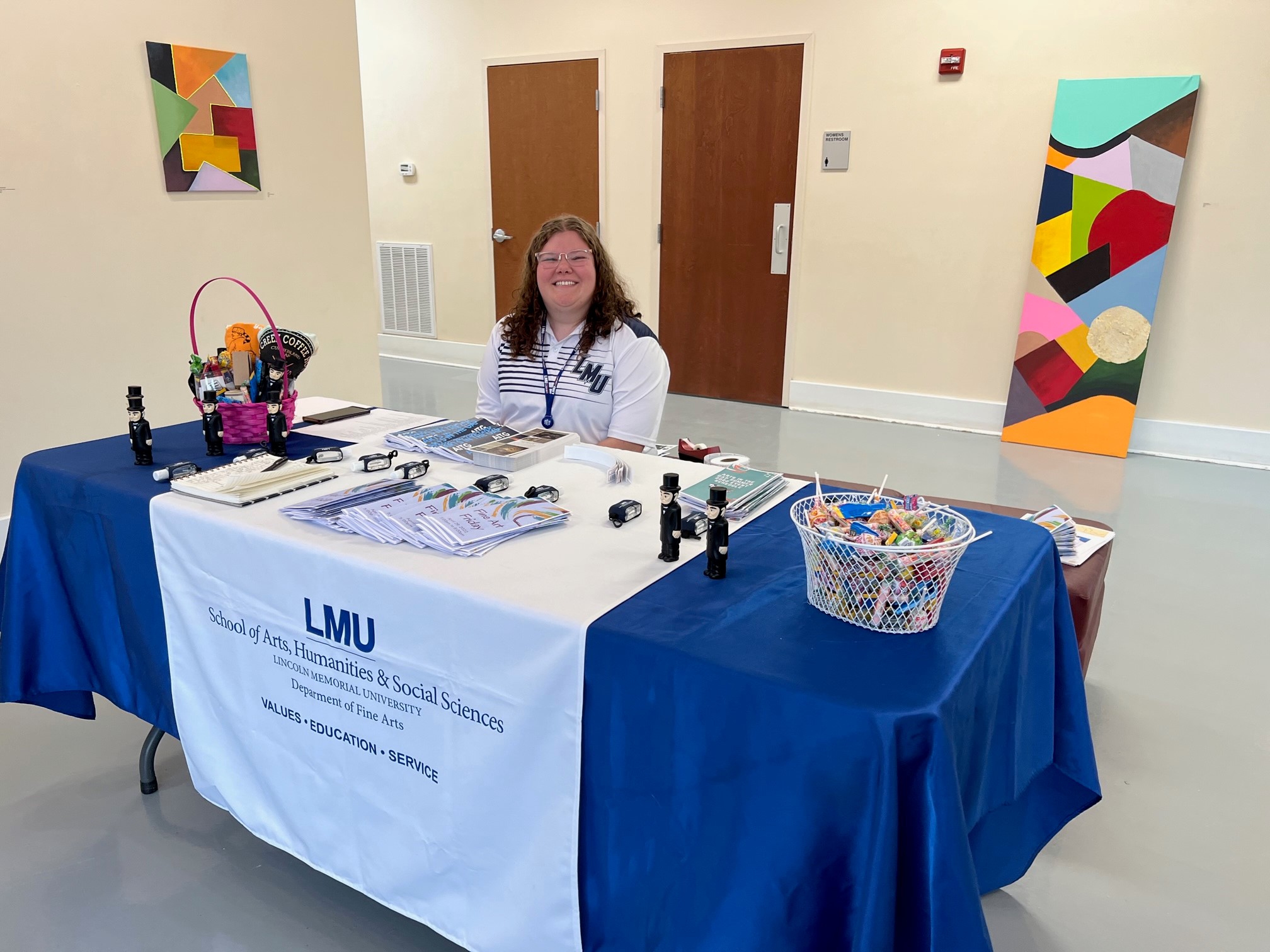 Grace, woman in her late 20s with curly brown hair in a polo. Sitting at a table surrounded by marketing materials for Arts in the Gap. 