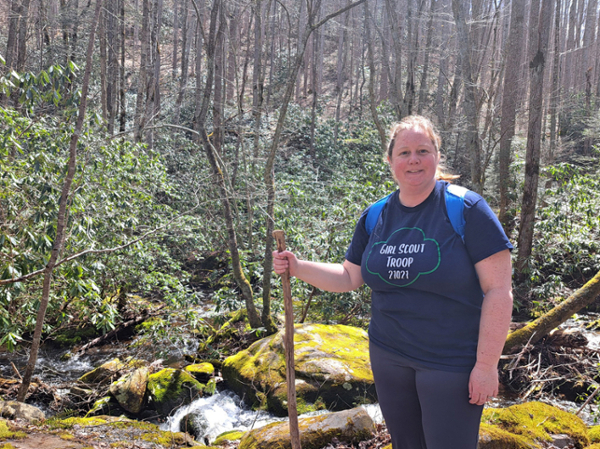 Smiling woman standing in a wooded area, holding a long walking stick. 