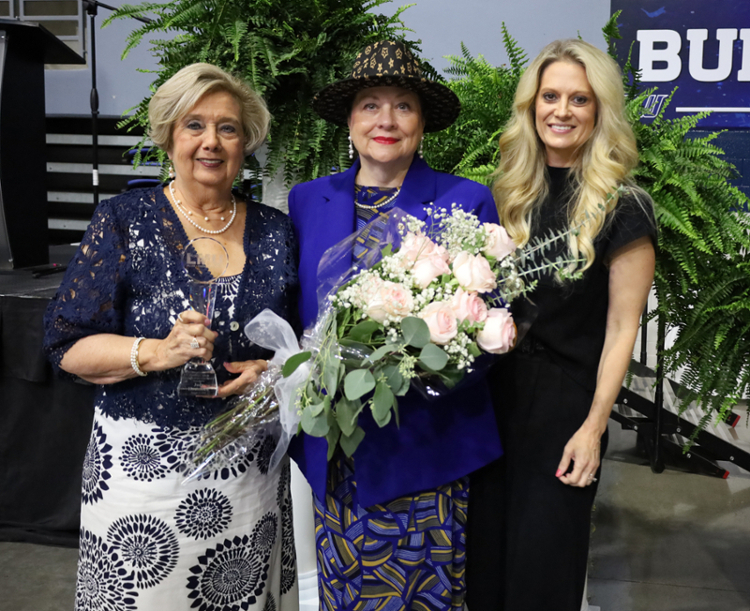 Three women standing in front of a stage. Left one is holding a glass award, center one is holding a bouquet of pink roses, right one is posing and smiling.